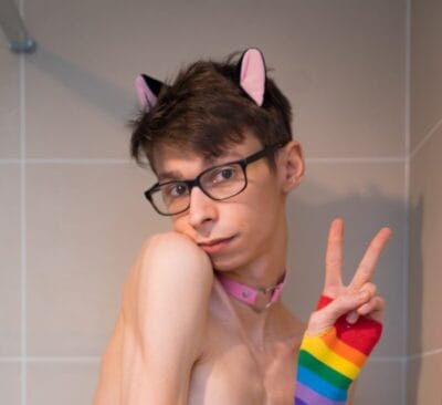 Person with short brown hair and glasses posing in a tiled shower, wearing pink cat-ear headband, pink collar, and a rainbow striped sleeve while making a peace sign with their hand.