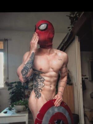Shirtless man wearing a Spider-Man mask poses indoors while holding a Captain America shield, showing tattoos on his torso and arm.