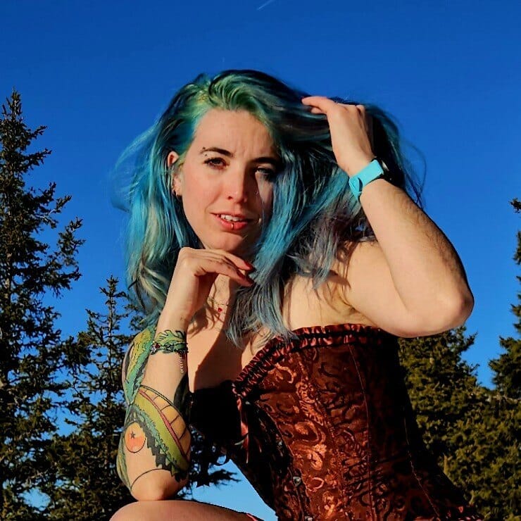 Woman with bright blue-green hair posing outdoors against a clear blue sky, wearing a burgundy corset and a turquoise wristband.