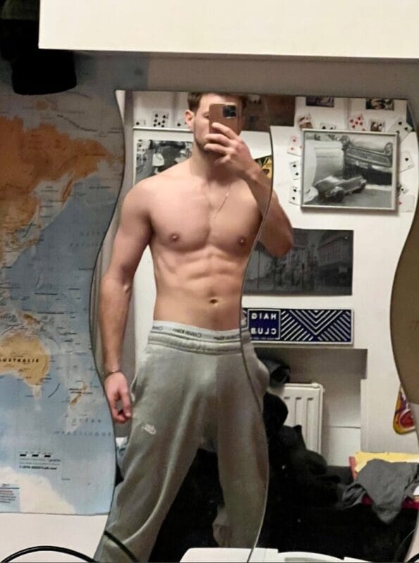 Young man taking a mirror selfie, shirtless, in a cluttered room wearing gray sweatpants, muscles visible.