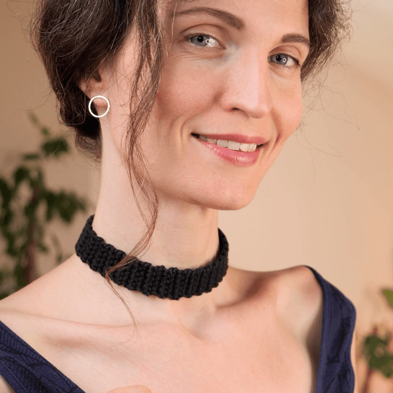 Close-up of a woman wearing a black crocheted choker and a white circular hoop earring, smiling subtly.
